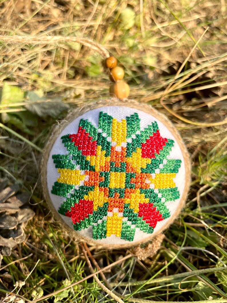 May include: A round, white ornament with a green, red, yellow, and orange geometric pattern embroidered on it. The ornament is made of fabric and has a burlap backing. It is hanging from a wooden bead.