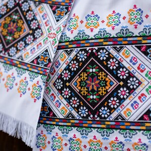 May include: White fabric with colorful embroidered floral patterns. The fabric has a fringe border.