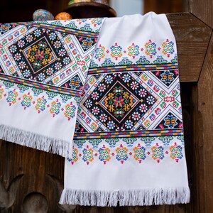 May include: Two white embroidered table runners with colorful geometric patterns and fringe edges. The runners are hanging on a wooden fence.