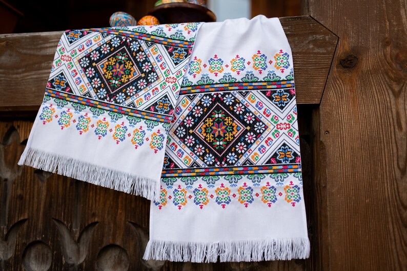 May include: Two white embroidered table runners with colorful geometric patterns and fringe edges. The runners are hanging on a wooden fence.