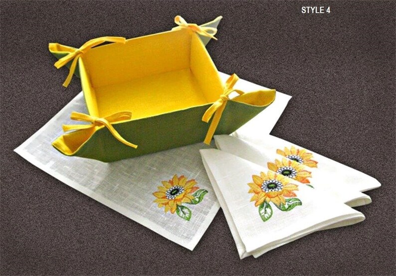 May include: A green and yellow felt bread basket with yellow ribbon ties. The basket is sitting on a white linen placemat with embroidered sunflowers. Three white linen napkins with embroidered sunflowers are folded next to the basket. The text "STYLE 4" is in the upper right corner of the image.