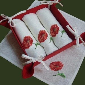May include: Set of three white linen napkins with red poppy embroidery, folded and displayed in a red felt basket with white ribbon ties.