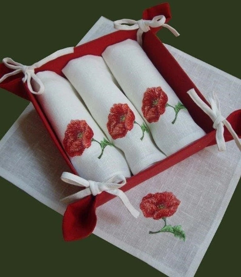 May include: Set of three white linen napkins with red poppy embroidery, folded and displayed in a red felt basket with white ribbon ties.