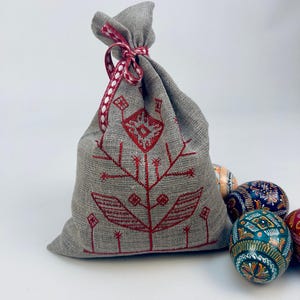 Puede incluir: Una bolsa de lino gris con un dise&ntilde;o floral rojo bordado, atada con una cinta roja y blanca. Varios huevos de Pascua decorados est&aacute;n esparcidos alrededor de la bolsa, con patrones intrincados y colores vibrantes.