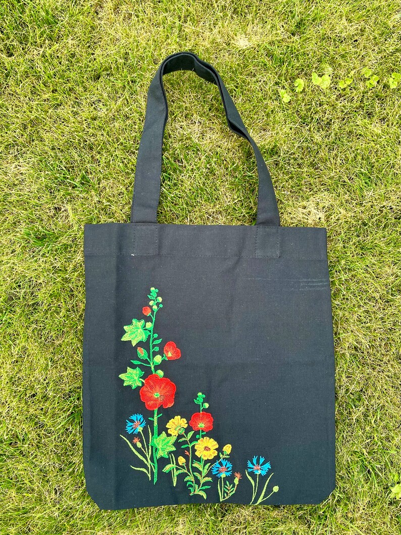 以下が含まれることがあります： 黒いキャンバス地のトートバッグに、花柄の刺繍が施されています。刺繍には、赤、黄、青の花と緑の葉が描かれています。