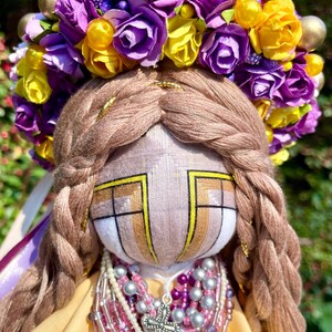 Puede incluir: Una mu&ntilde;eca hecha a mano con un vestido amarillo, una corona de flores moradas y amarillas y un collar de cuentas con un colgante de cruz de plata.