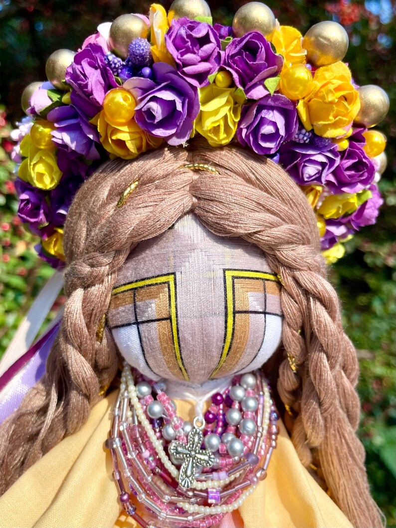 Puede incluir: Una mu&ntilde;eca hecha a mano con un vestido amarillo, una corona de flores moradas y amarillas y un collar de cuentas con un colgante de cruz de plata.