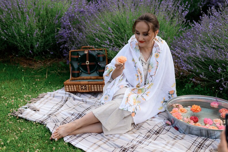 Pode incluir: Uma mulher est&aacute; sentada em uma manta xadrez em um campo de lavanda, segurando uma flor rosa. Uma cesta de piquenique com uma tampa verde est&aacute; ao lado dela. Uma tigela de metal cheia de &aacute;gua e flores rosas est&aacute; na frente dela.