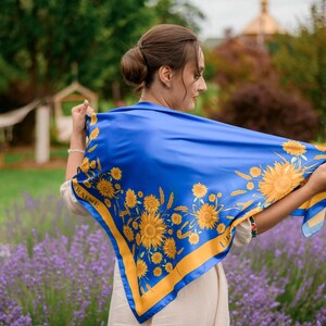 May include: A blue and yellow silk scarf with a sunflower design. The scarf has a blue background with yellow sunflowers and wheat stalks. The scarf is being held up by a person in a garden. The text "Style 2" is in the top right corner.