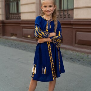Pu&ograve; includere: Una bambina indossa un vestito blu con ricami gialli. L'abito presenta maniche lunghe con motivi a forma di grano e nappe decorative. Indossa scarpe nere e si trova su una superficie pavimentata.