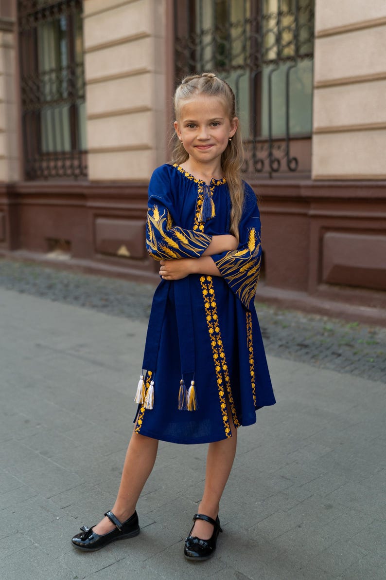 Pu&ograve; includere: Una bambina indossa un vestito blu con ricami gialli. L'abito presenta maniche lunghe con motivi a forma di grano e nappe decorative. Indossa scarpe nere e si trova su una superficie pavimentata.