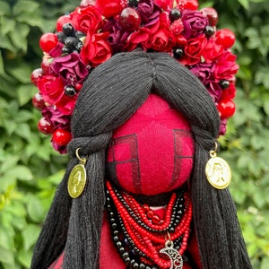 Puede incluir: Una mu&ntilde;eca de tela roja con cabello negro que lleva una corona de flores rojas, un collar de cuentas rojas y negras y pendientes de oro. La mu&ntilde;eca est&aacute; vestida con un vestido rojo con un borde de encaje negro.