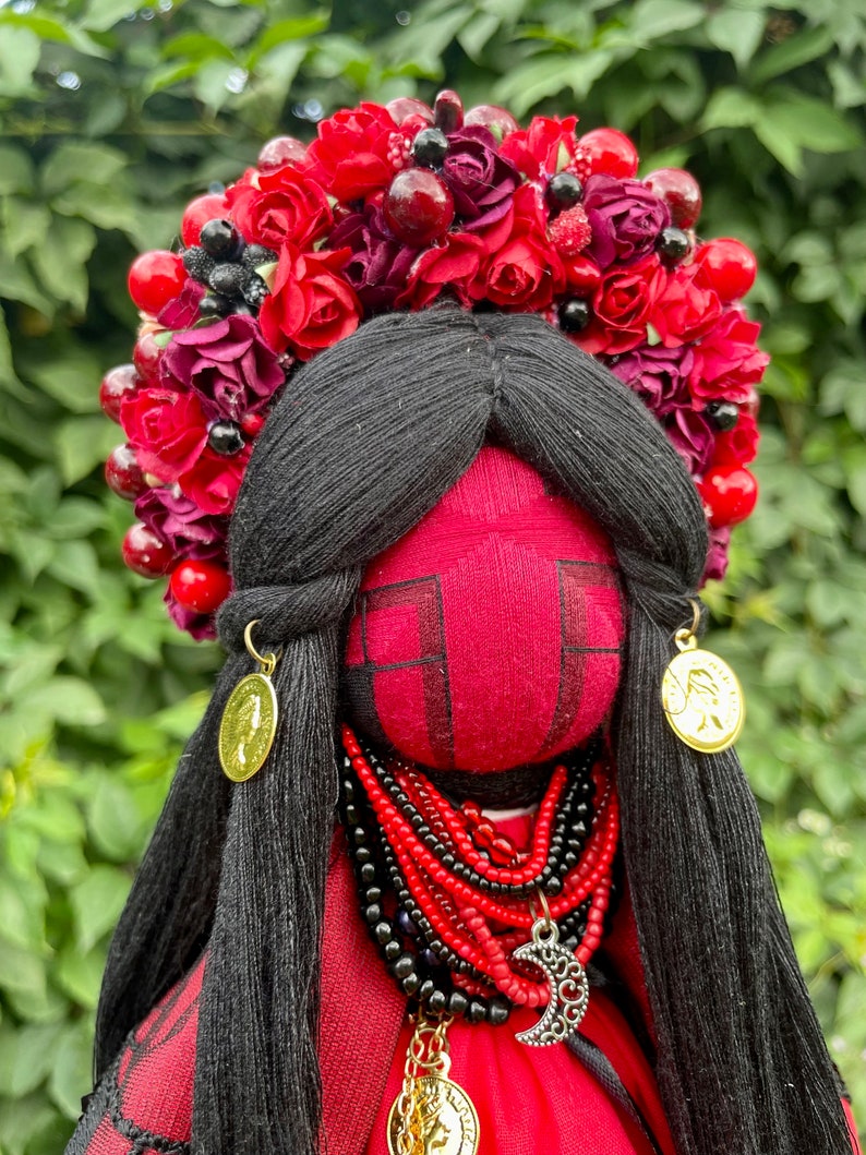 Puede incluir: Una mu&ntilde;eca de tela roja con cabello negro que lleva una corona de flores rojas, un collar de cuentas rojas y negras y pendientes de oro. La mu&ntilde;eca est&aacute; vestida con un vestido rojo con un borde de encaje negro.