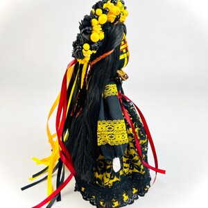 K&ouml;nnte beinhalten: Handgefertigte Puppe mit langem, schwarzem Haar und einem Blumenkranz. Die Puppe tr&auml;gt ein schwarzes Kleid mit gelben Blumenmustern und Spitzenbesatz. Rote, gelbe und schwarze B&auml;nder fallen von der Krone. Die Puppe ist ein traditionelles Volkskunstwerk.