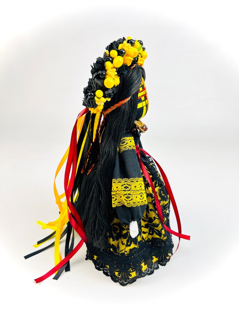 K&ouml;nnte beinhalten: Handgefertigte Puppe mit langem, schwarzem Haar und einem Blumenkranz. Die Puppe tr&auml;gt ein schwarzes Kleid mit gelben Blumenmustern und Spitzenbesatz. Rote, gelbe und schwarze B&auml;nder fallen von der Krone. Die Puppe ist ein traditionelles Volkskunstwerk.