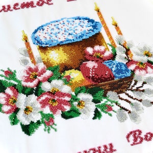 K&ouml;nnte beinhalten: Besticktes wei&szlig;es Tuch mit einem farbenfrohen Osterdesign. Das Design umfasst einen Korb mit dekorierten Eiern, einen Kuchen, Kerzen und florale Akzente. Der Text "Christus ist auferstanden" ist rot gestickt.