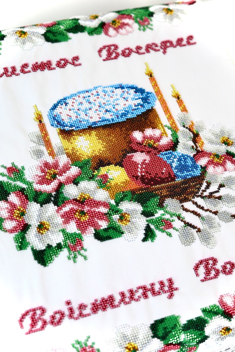 K&ouml;nnte beinhalten: Besticktes wei&szlig;es Tuch mit einem farbenfrohen Osterdesign. Das Design umfasst einen Korb mit dekorierten Eiern, einen Kuchen, Kerzen und florale Akzente. Der Text "Christus ist auferstanden" ist rot gestickt.