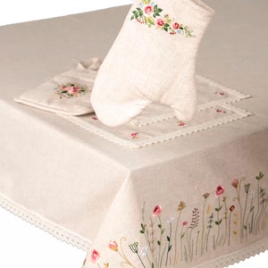 May include: A beige linen oven mitt with floral embroidery, held above a matching placemat and tablecloth. The tablecloth features embroidered flowers and a lace trim. The set is designed for kitchen use.