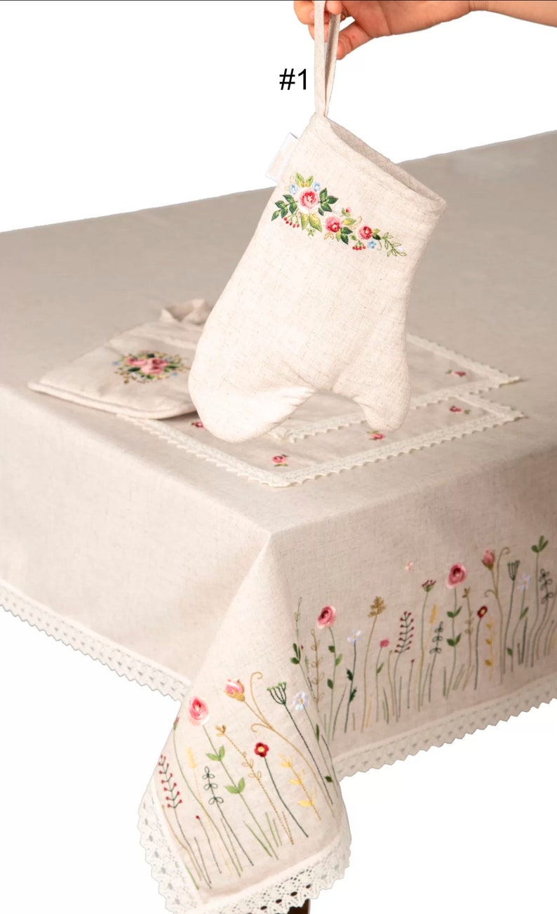 May include: A beige linen oven mitt with floral embroidery, held above a matching placemat and tablecloth. The tablecloth features embroidered flowers and a lace trim. The set is designed for kitchen use.