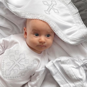 May include: A white baby outfit with a cross embroidered on the front. The outfit includes a long-sleeved shirt, pants, and a hat. The baby is wearing the outfit and is lying on a white blanket.
