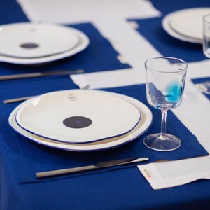 May include: A table setting with white plates featuring a dark purple center and a blue rim, set on a deep blue tablecloth. Clear wine glasses and silverware complete the elegant dining arrangement. White placemats add contrast.