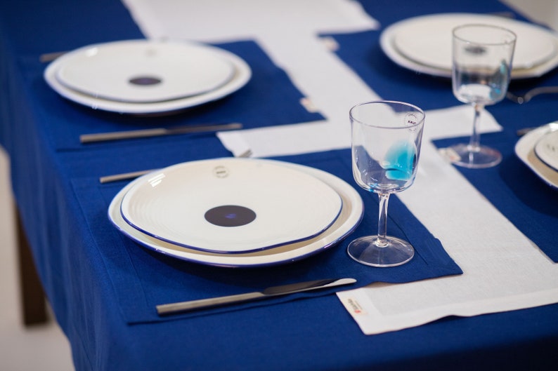 May include: A table setting with white plates featuring a dark purple center and a blue rim, set on a deep blue tablecloth. Clear wine glasses and silverware complete the elegant dining arrangement. White placemats add contrast.