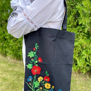 以下が含まれることがあります： 黒いトートバッグに花柄の刺繍が施されています。刺繍には赤、黄、青の花と緑の葉が描かれています。