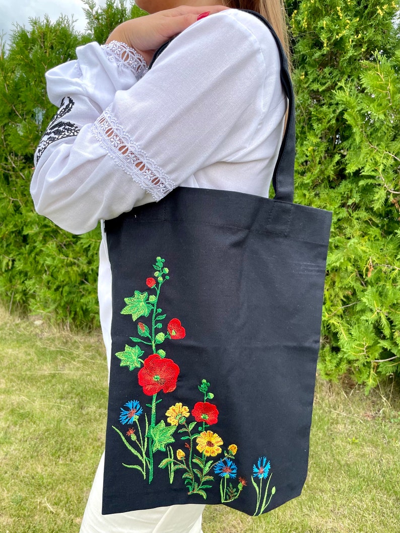 以下が含まれることがあります： 黒いトートバッグに花柄の刺繍が施されています。刺繍には赤、黄、青の花と緑の葉が描かれています。