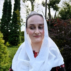 May include: A woman wearing a white headscarf with a lace trim and a black embroidered blouse with red and white floral patterns. The background features green trees and foliage.