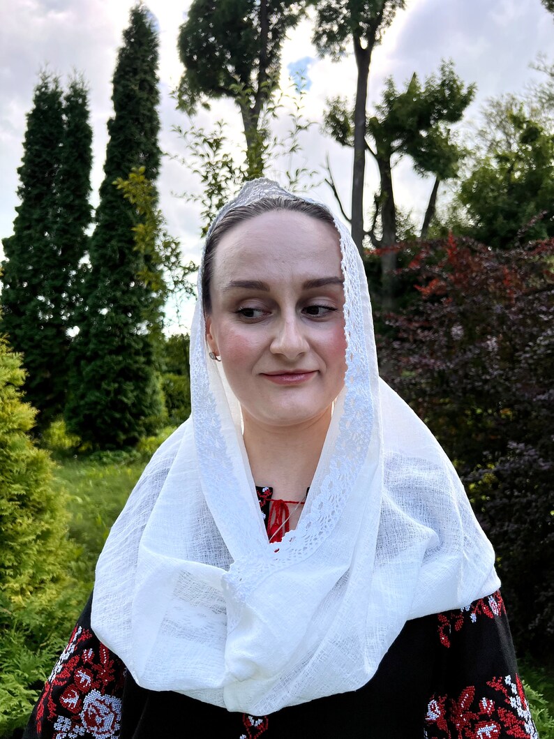 May include: A woman wearing a white headscarf with a lace trim and a black embroidered blouse with red and white floral patterns. The background features green trees and foliage.