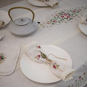 May include: A white teapot with a gold handle sits on a white linen tablecloth with embroidered floral designs. A white plate with a folded linen napkin sits on the table.