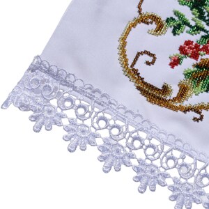 K&ouml;nnte beinhalten: Wei&szlig;er Stoff mit zarter, floraler Spitzenborte. Die Ecke des Stoffes zeigt ein gesticktes Design mit goldenen, gr&uuml;nen und roten Akzenten. Die Stickerei ist detailliert und verziert und verleiht einen Hauch von Eleganz.