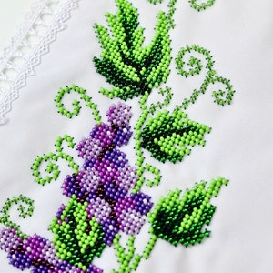Puede incluir: Primer plano de tela blanca con bordado de cuentas. El dise&ntilde;o presenta un racimo de uvas moradas y hojas verdes con un ribete de encaje blanco. El bordado es detallado y vibrante.