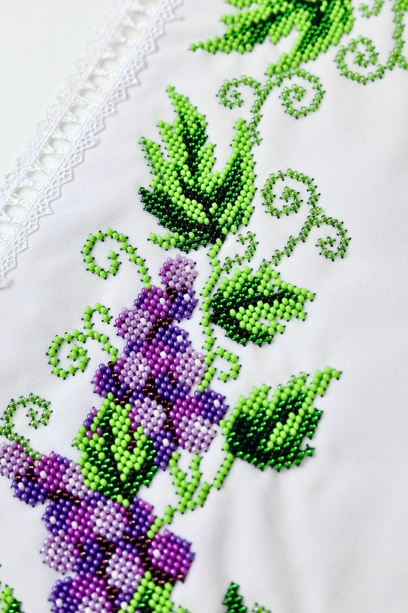 Puede incluir: Primer plano de tela blanca con bordado de cuentas. El dise&ntilde;o presenta un racimo de uvas moradas y hojas verdes con un ribete de encaje blanco. El bordado es detallado y vibrante.