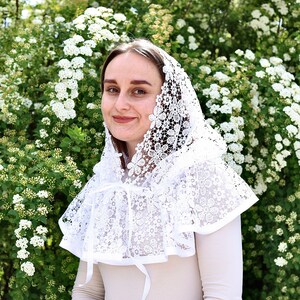 May include: A white lace head covering with a scalloped edge and floral pattern. The head covering is worn by a person with brown hair and is tied at the neck with a white ribbon.