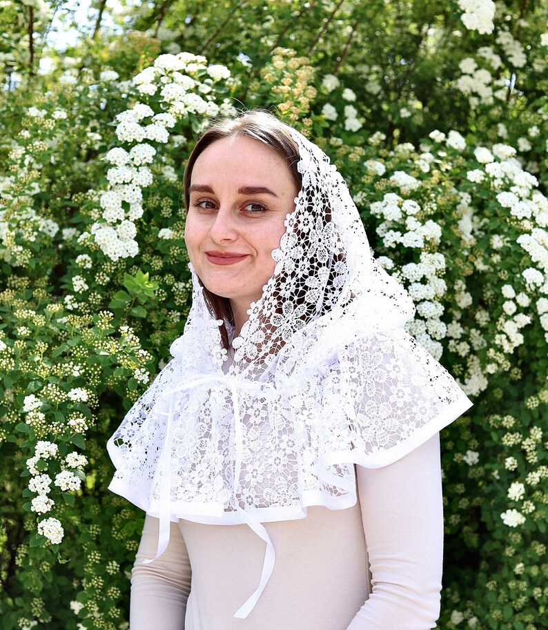 May include: A white lace head covering with a scalloped edge and floral pattern. The head covering is worn by a person with brown hair and is tied at the neck with a white ribbon.