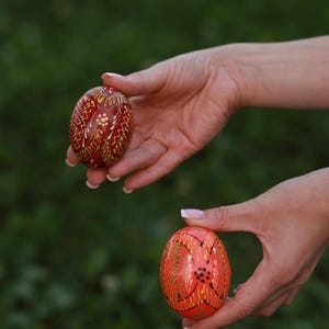 K&ouml;nnte beinhalten: Zwei kunstvoll bemalte Ostereier, eines rot mit goldenen und wei&szlig;en Mustern und eines orange mit goldenen und schwarzen Mustern, werden in den H&auml;nden einer Person gehalten.