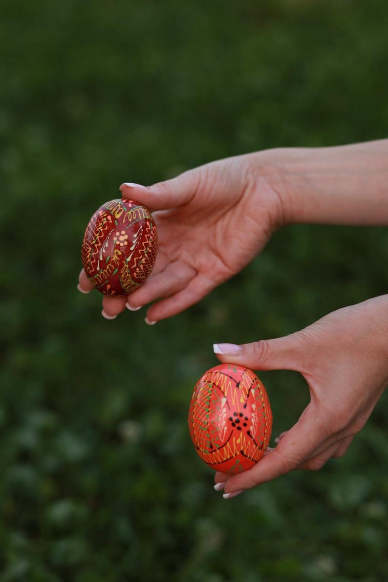 K&ouml;nnte beinhalten: Zwei kunstvoll bemalte Ostereier, eines rot mit goldenen und wei&szlig;en Mustern und eines orange mit goldenen und schwarzen Mustern, werden in den H&auml;nden einer Person gehalten.