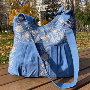 Peut inclure: Un sac cabas en toile bleu clair avec des motifs floraux brod&eacute;s blancs. Le sac a une longue bandouli&egrave;re et deux anneaux m&eacute;talliques en haut.