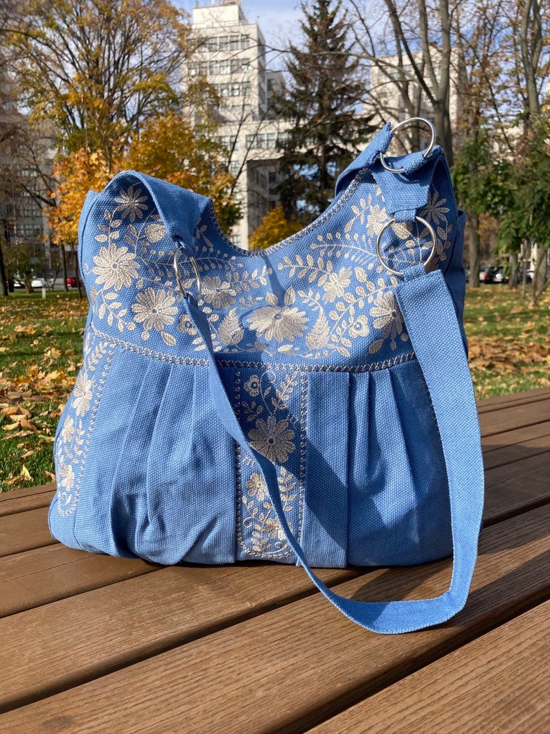 Peut inclure: Un sac cabas en toile bleu clair avec des motifs floraux brod&eacute;s blancs. Le sac a une longue bandouli&egrave;re et deux anneaux m&eacute;talliques en haut.