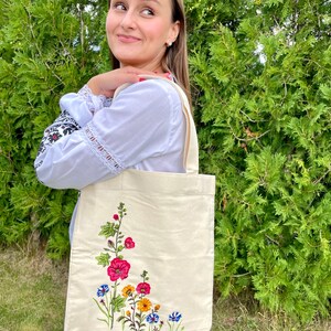 以下が含まれることがあります： 白いキャンバス地のトートバッグに、花柄の刺繍が施されています。刺繍には、ピンク、赤、黄色、青の花と緑の葉が描かれています。