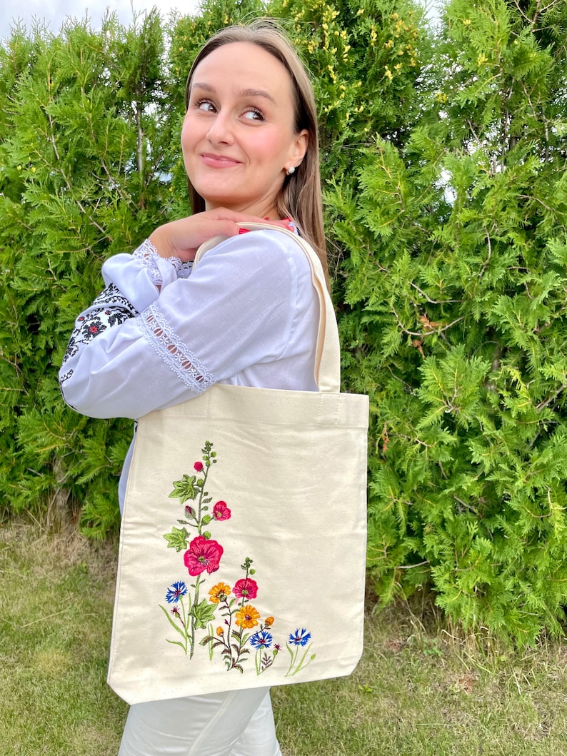 以下が含まれることがあります： 白いキャンバス地のトートバッグに、花柄の刺繍が施されています。刺繍には、ピンク、赤、黄色、青の花と緑の葉が描かれています。