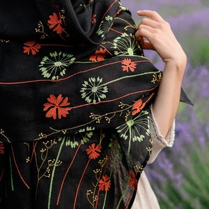May include: Black scarf with embroidered floral design in orange, green, and yellow. The scarf is draped over the shoulders. The background is a field of purple flowers.