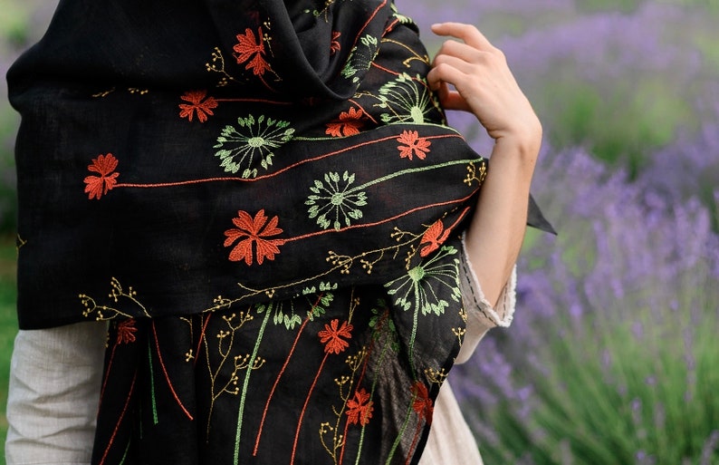 May include: Black scarf with embroidered floral design in orange, green, and yellow. The scarf is draped over the shoulders. The background is a field of purple flowers.