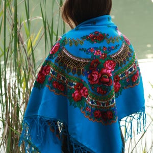May include: A vibrant blue shawl with a floral pattern in red, pink, and green. The shawl features a decorative border and fringe along the edges. The design is reminiscent of traditional folk art.