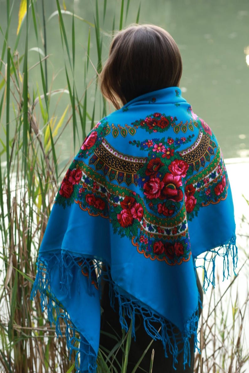 May include: A vibrant blue shawl with a floral pattern in red, pink, and green. The shawl features a decorative border and fringe along the edges. The design is reminiscent of traditional folk art.