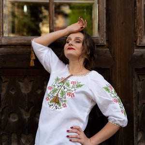 May include: A woman wearing a white embroidered blouse with a floral design and brown tassels. She is standing in front of a wooden door with a window.