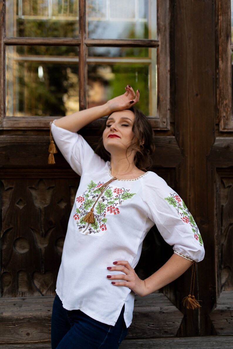 May include: A woman wearing a white embroidered blouse with a floral design and brown tassels. She is standing in front of a wooden door with a window.