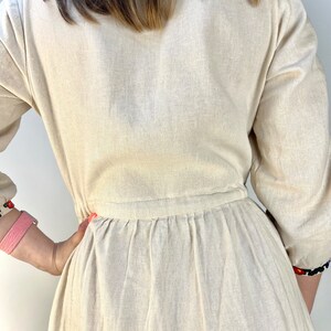 May include: Beige linen dress with long sleeves and a gathered waist. The dress features embroidered details on the sleeves and neckline. The dress is a neutral color and has a simple, elegant design.