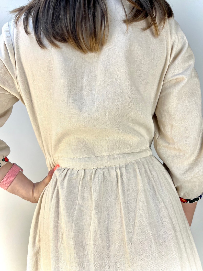 May include: Beige linen dress with long sleeves and a gathered waist. The dress features embroidered details on the sleeves and neckline. The dress is a neutral color and has a simple, elegant design.